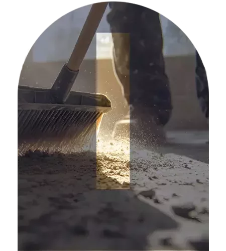 A close-up of a broom sweeping dust and debris across a floor, with sunlight highlighting the particles in the air. The image is overlaid with a large translucent letter "I" in the centre.